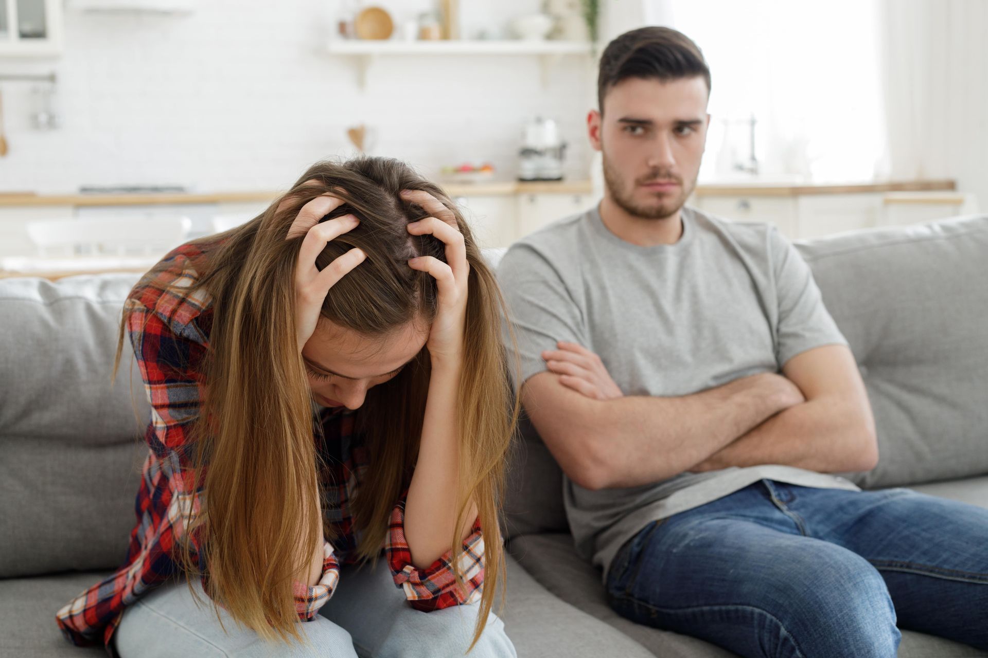 Woman with head in hands, distraught, beside a man with arms crossed, looking stern; both on a sofa.