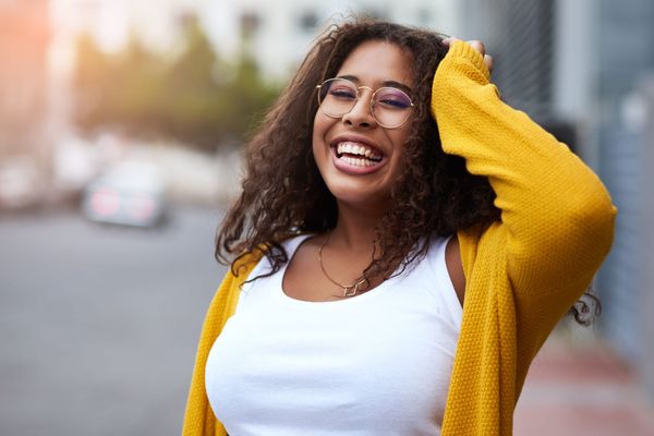 Woman in yellow sweater laughs outdoors, touches her curly hair.