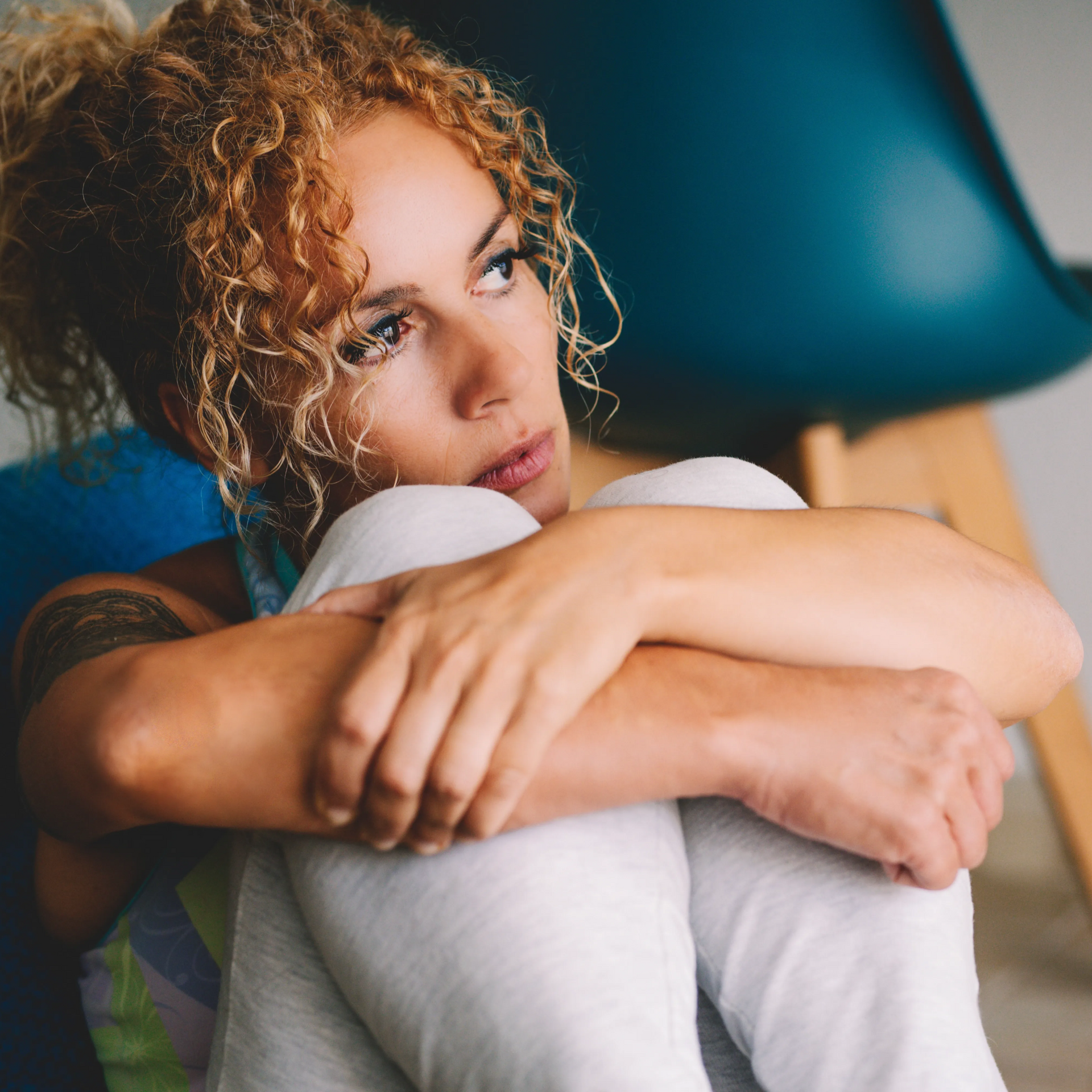Woman with curly hair sits hugging her knees, looking contemplative. Leaning against a blue chair.