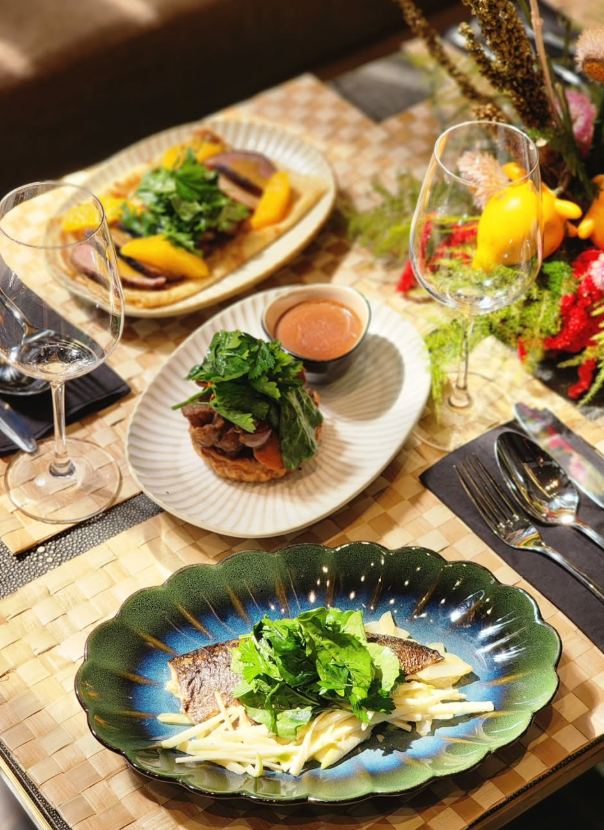 Three plates of food on a table set for dining, with greenery and wine glasses.