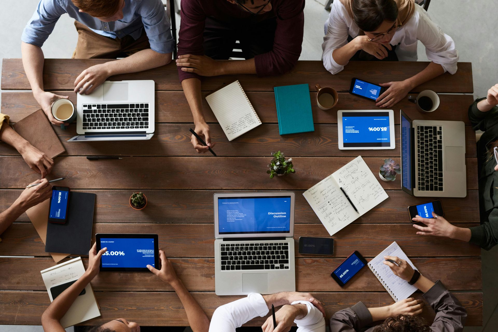 Group of people at a wooden table with laptops, tablets, phones, and notebooks, appearing to collaborate.