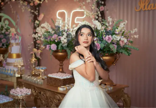 Donna in abito bianco in posa davanti a un tavolo di dolci floreale con decorazioni al neon numero 16 e rosa.
