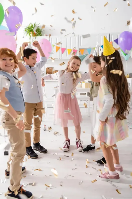Bambini con cappellini da festa che festeggiano con coriandoli in una stanza decorata a festa.