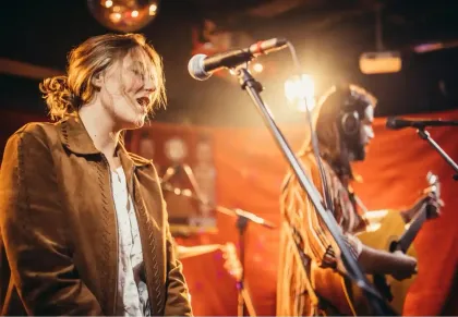 Il gruppo si esibisce dal vivo sul palco, con il cantante al microfono e il chitarrista immersi in una calda luce arancione.