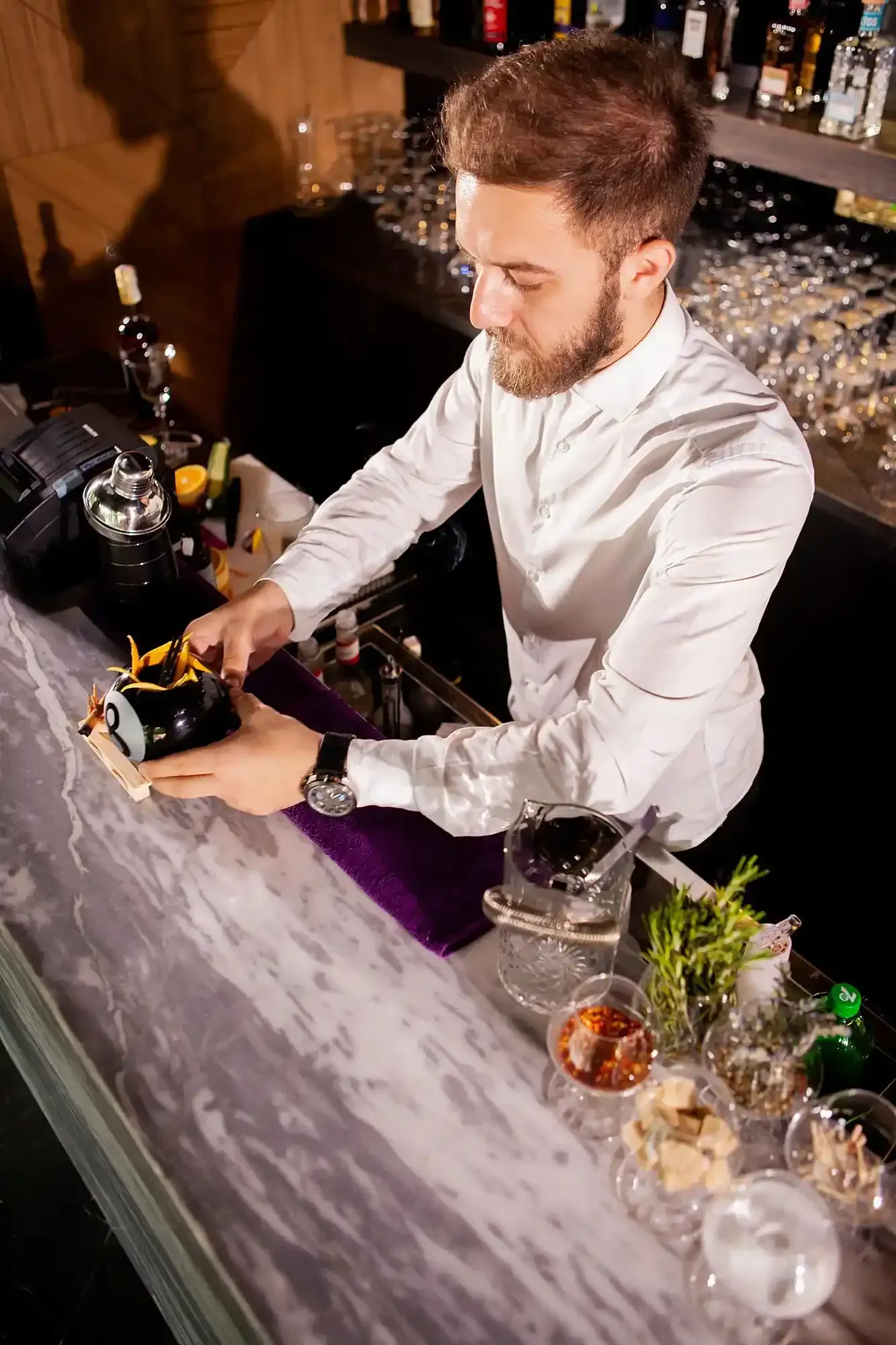 Un barista in camicia bianca prepara un drink dietro un bancone di marmo, utilizzando utensili e bicchieri da cocktail.