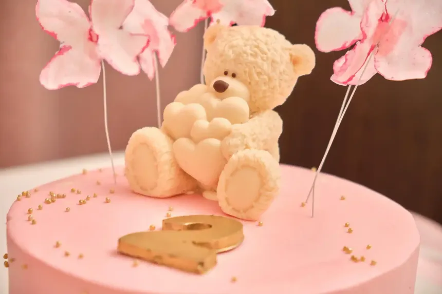 Orsetto di peluche su una torta rosa con fiori pastello e zuccherini dorati.