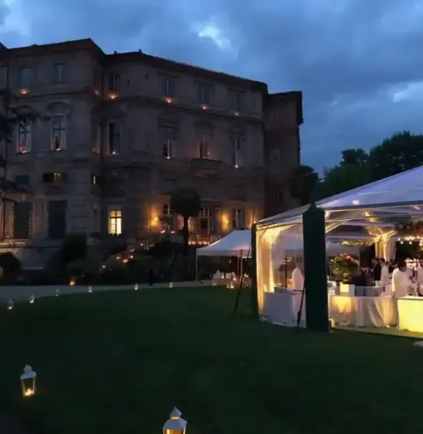 Evento serale all'aperto accanto a una villa illuminata, con un tendone bianco sul prato sotto un cielo azzurro nuvoloso.