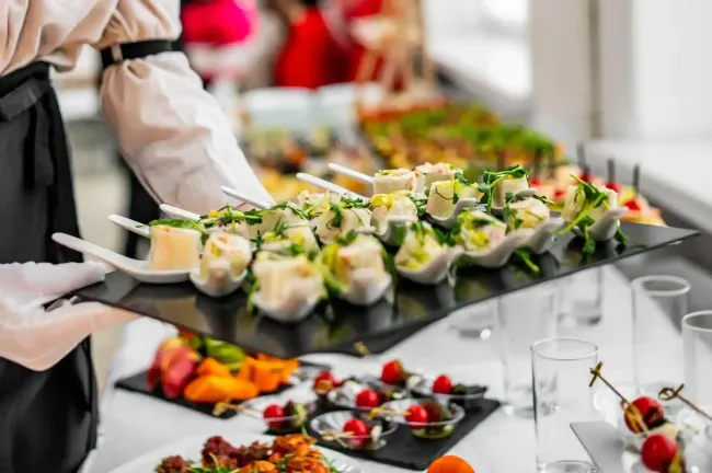 Un addetto al catering serve antipasti su un vassoio a un tavolo da buffet con frutta, dessert e bevande.