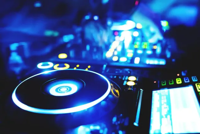 Primo piano di un mixer da DJ e di un giradischi che brillano di luce blu e bianca in un locale notturno buio.