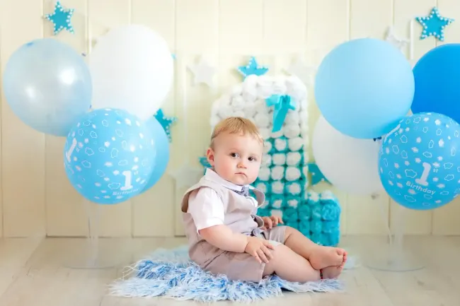 Un bambino seduto su una coperta blu tra palloncini blu e bianchi in una stanza decorata con colori vivaci.