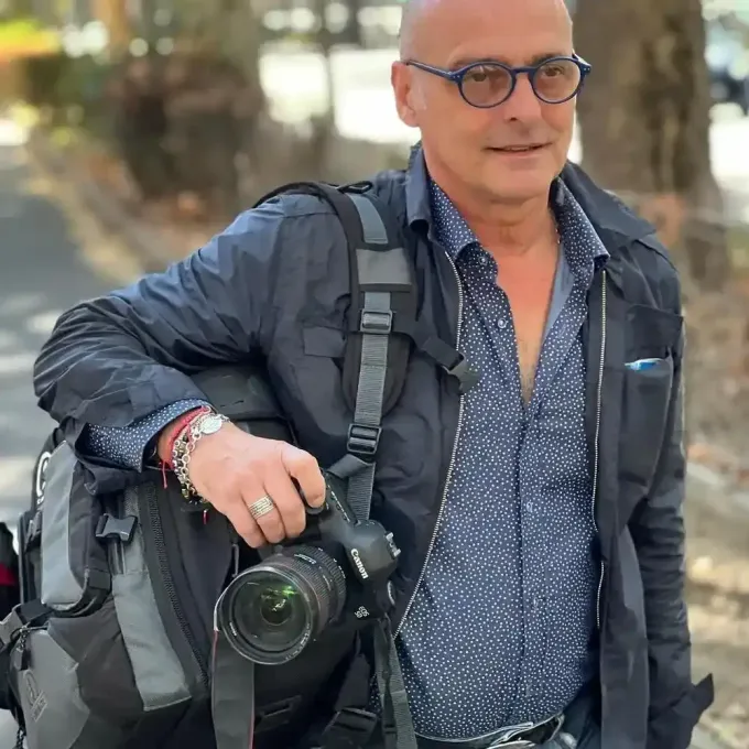 Uomo con gli occhiali che tiene in mano una macchina fotografica, con indosso una giacca scura, all'aperto su un sentiero alberato.