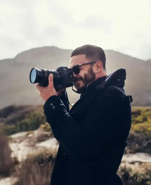 Una persona con giacca nera e occhiali da sole fotografa un paesaggio montano con una macchina fotografica.