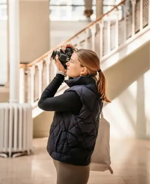 Una persona con un gilet nero scatta foto in casa accanto a una scala e a un termosifone.