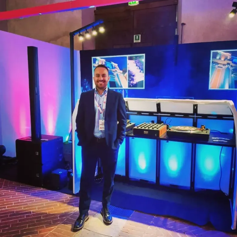 Un uomo in piedi davanti a uno stand fieristico illuminato di blu, con schermi e tavoli espositivi sullo sfondo.