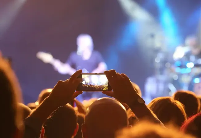La folla riprende un concerto dal vivo con uno smartphone, le luci del palco brillano di blu e arancione.