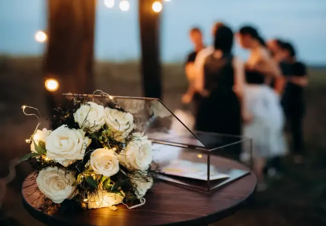 Un mazzo di rose bianche su un tavolino con gli ospiti sfocati durante un evento serale all'aperto.