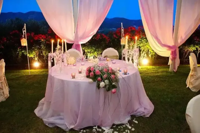 Romantico tavolo nuziale all'aperto con drappi bianchi, candele e centrotavola floreale rosa al crepuscolo