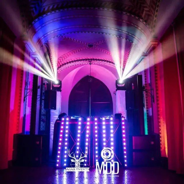 Palco illuminato di viola in una sala riccamente decorata con una cabina DJ a LED luminosa e ampi fasci di riflettori