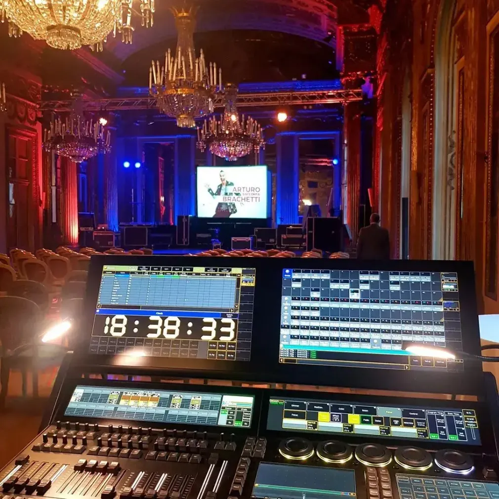 Cabina di controllo con vista su una sala da ballo illuminata di rosso, completa di schermo scenico, lampadari e console di missaggio audiovisivo.