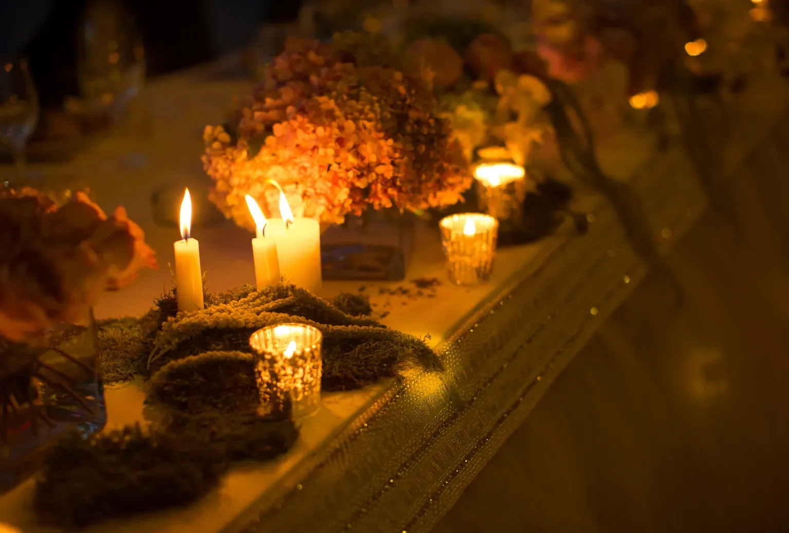 Candele luminose su un tavolo con composizioni floreali in un'atmosfera soffusa e calda.
