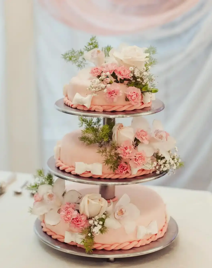 Torta a tre piani con decorazioni floreali dai colori pastello, su un tavolo bianco con fiori rosa e bianchi.