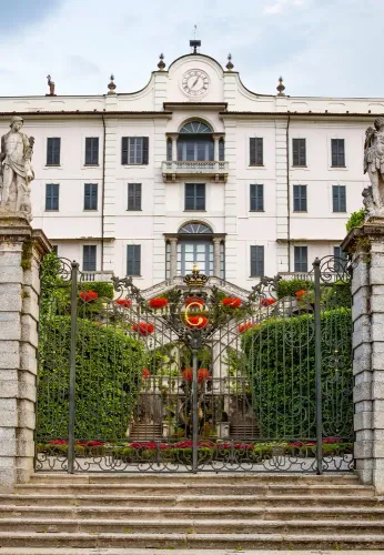 Edificio bianco riccamente decorato con orologio e balcone, situato dietro un cancello in ferro e con ingresso al giardino.
