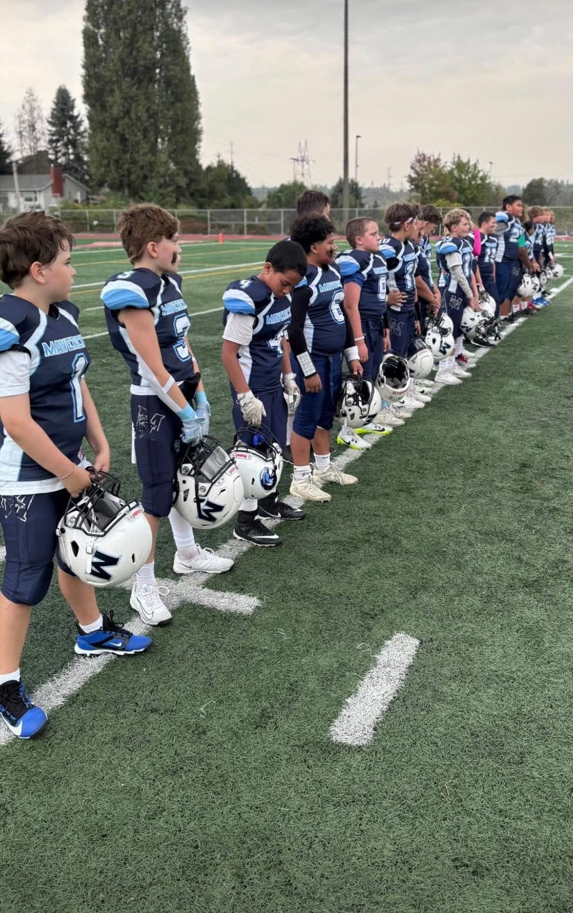 Youth football team in blue uniforms lined up on a field, holding helmets before practice.