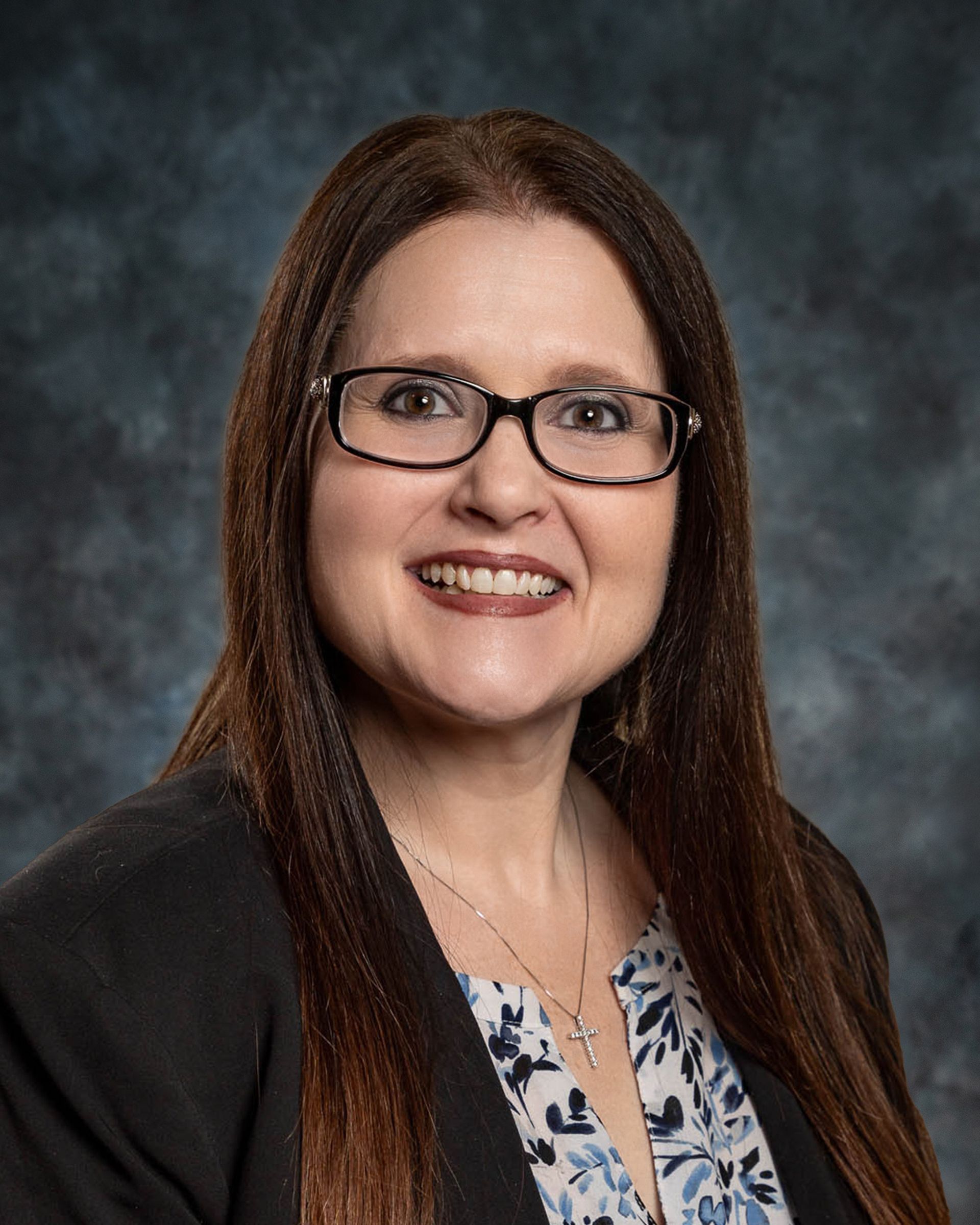 Woman with long brown hair wearing glasses and a black blazer smiles at the camera.