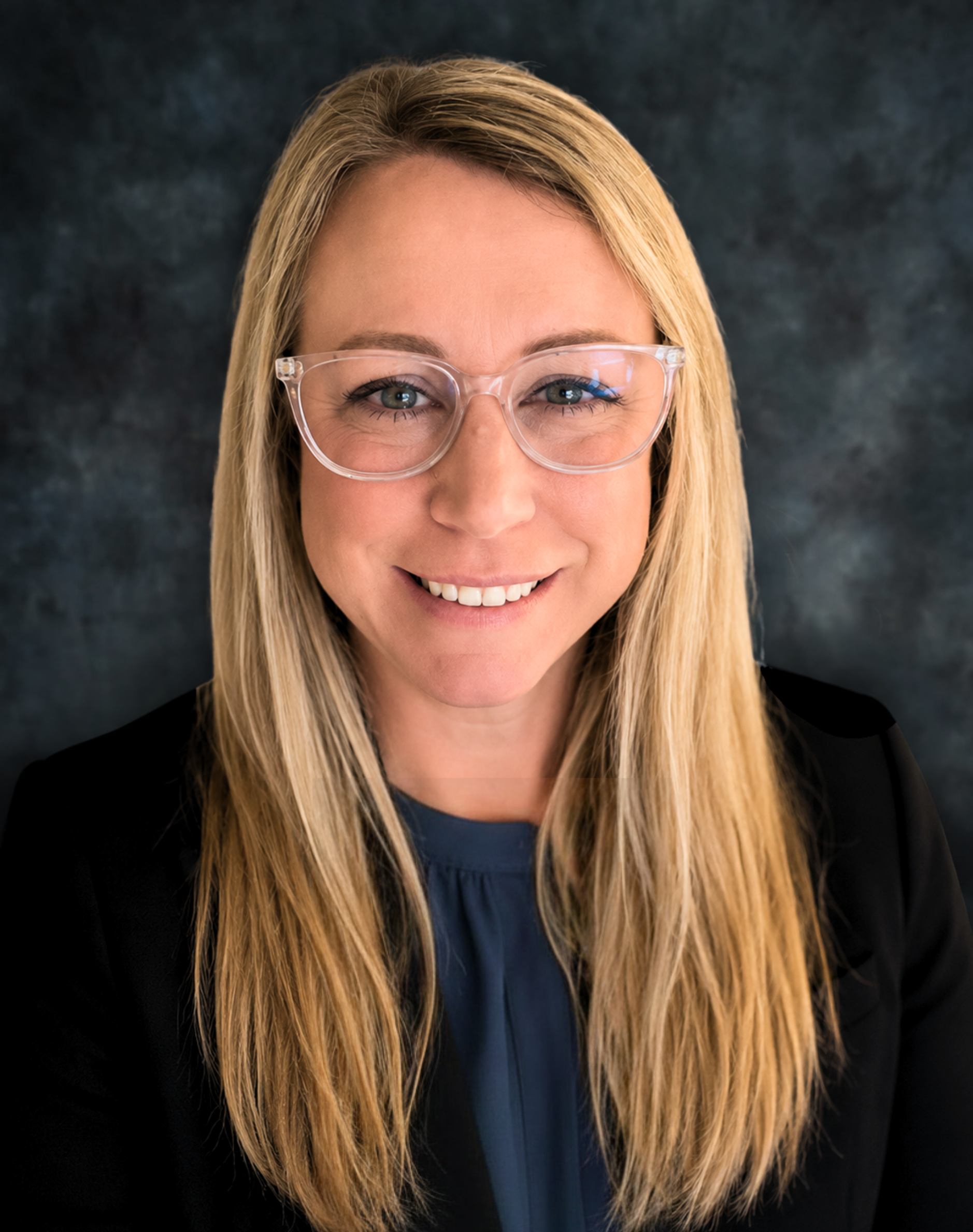 Woman with blonde hair wearing a dark blazer and smiling in front of a blue background.