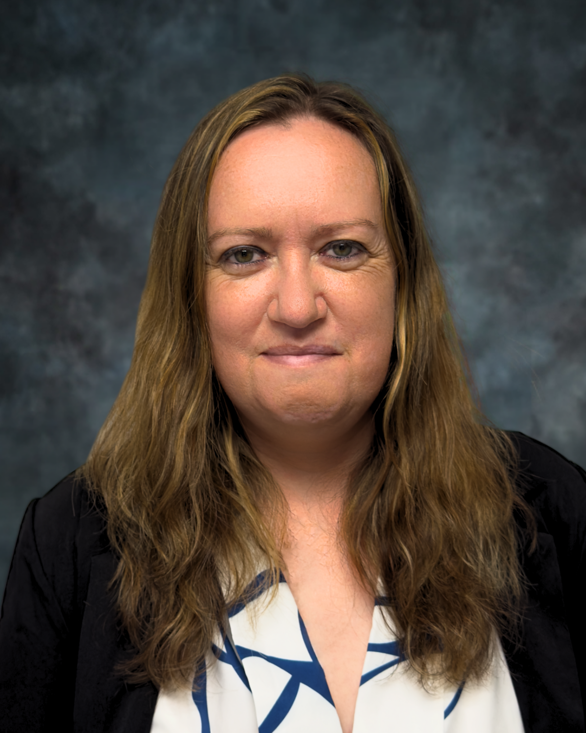 A person with long brown hair, wearing a black blazer over a white top with a blue pattern, against a mottled gray wall.