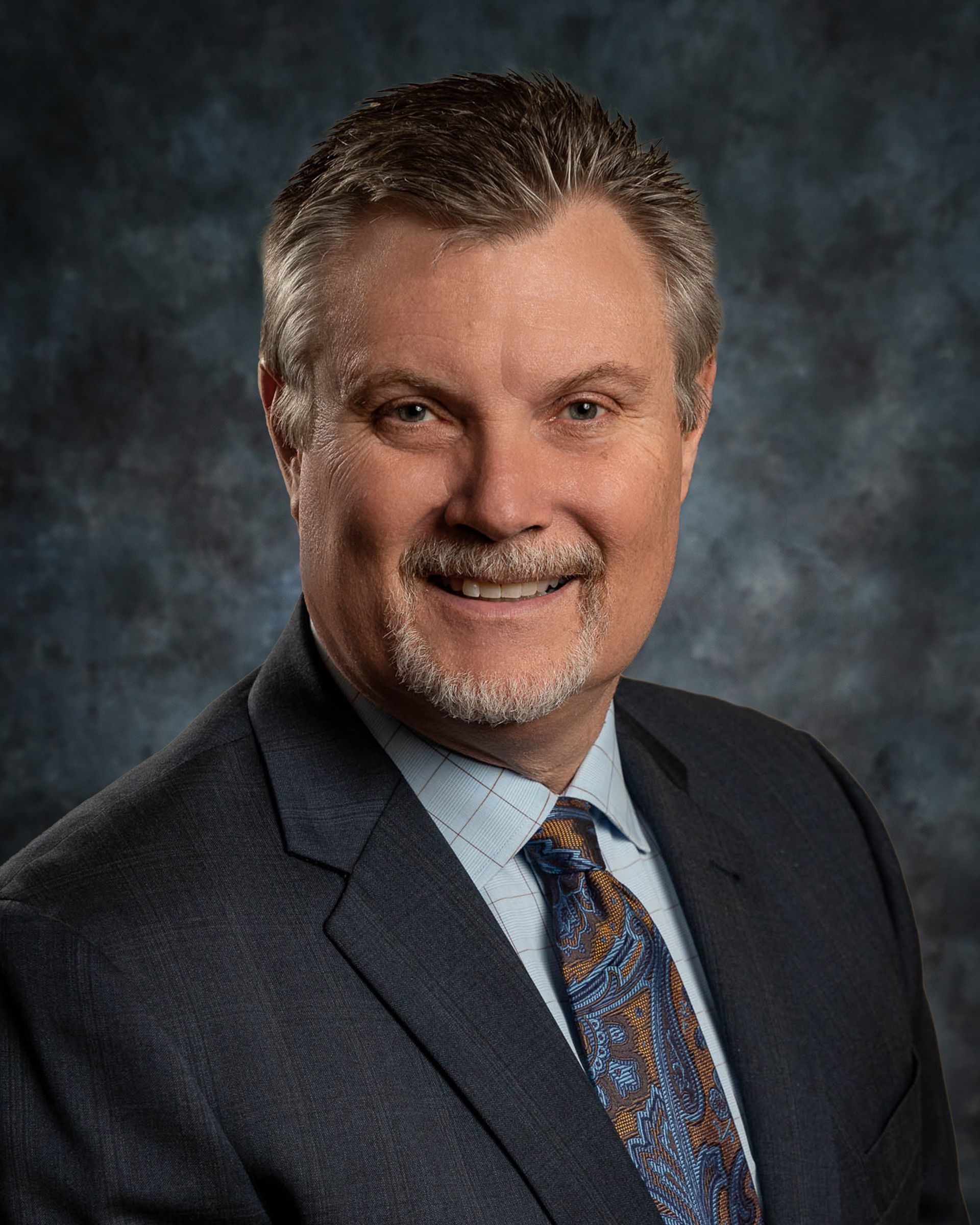 Man in a suit smiles at the camera, with a dark background.