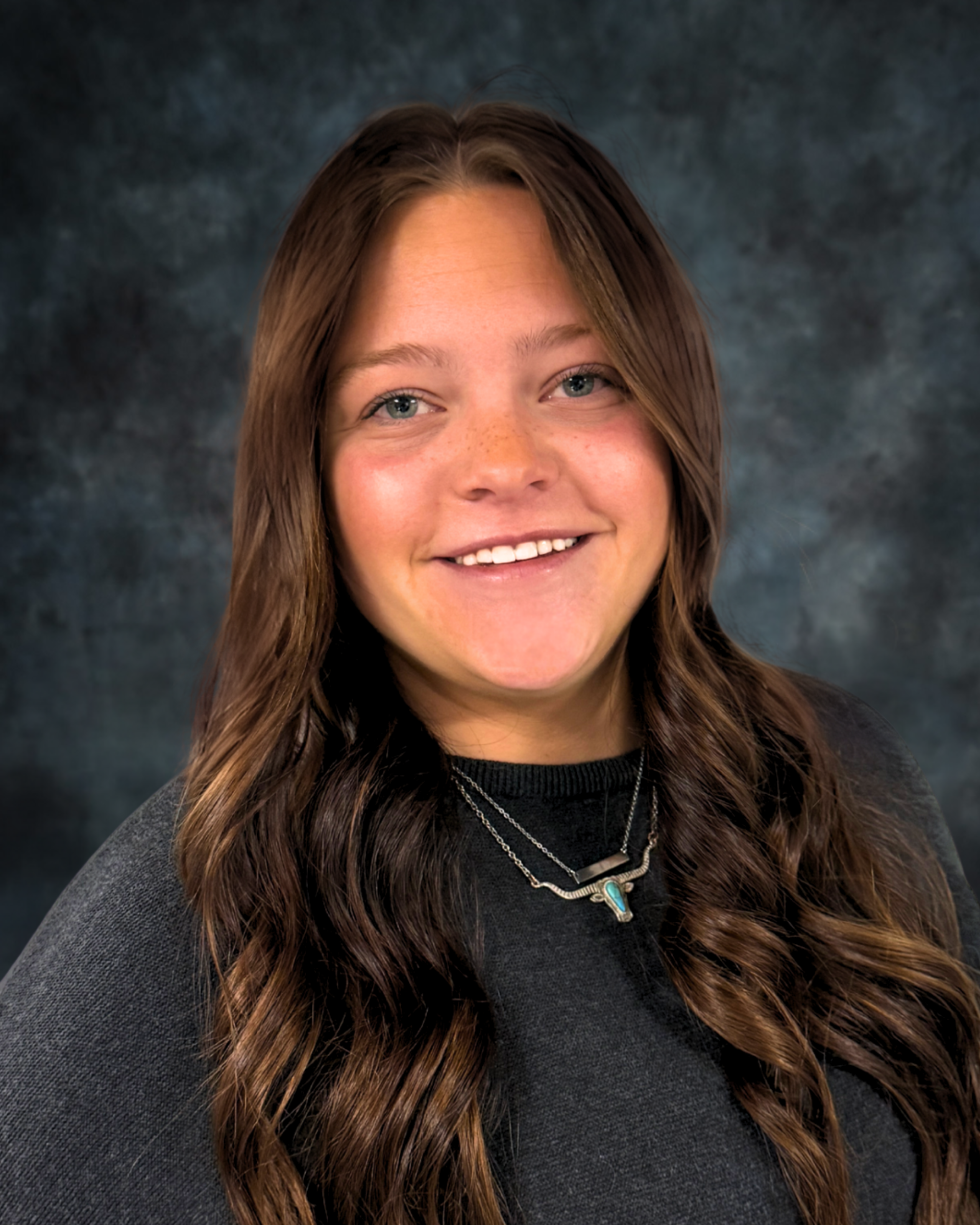 A smiling person with long, wavy brown hair wearing a dark sweater and a silver necklace against a dark mottled backdrop.