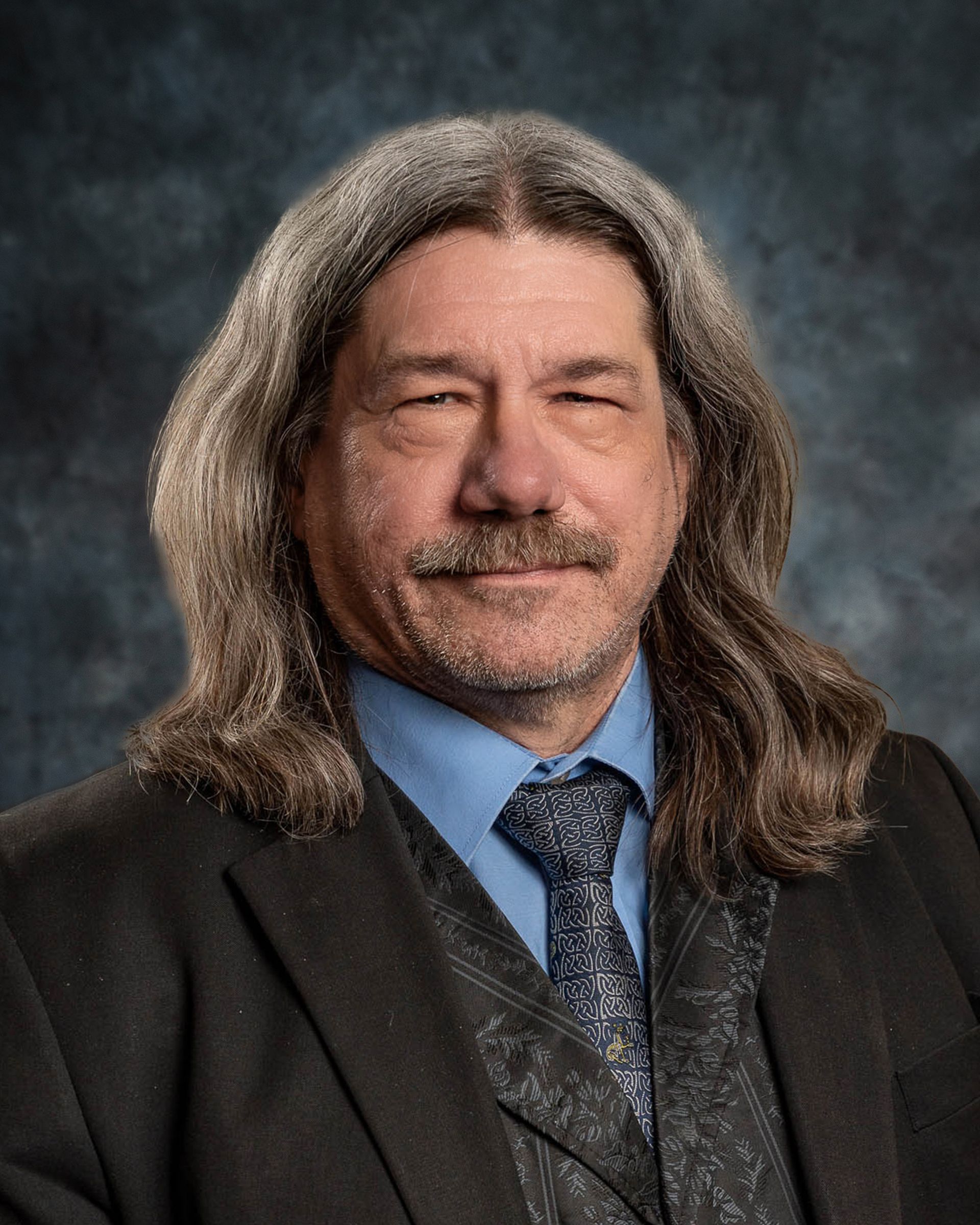 Man with long gray hair and mustache, wearing a suit and tie, smiling at the camera.