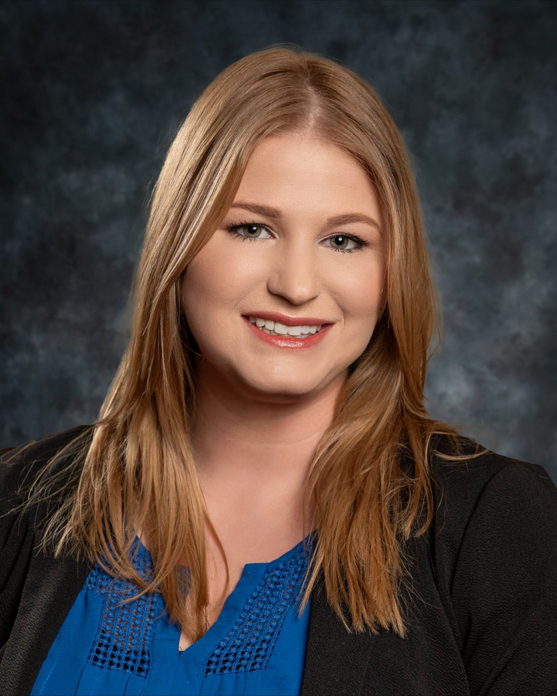 Blond woman smiling, wearing a blue shirt and black jacket. Against a dark, patterned background.