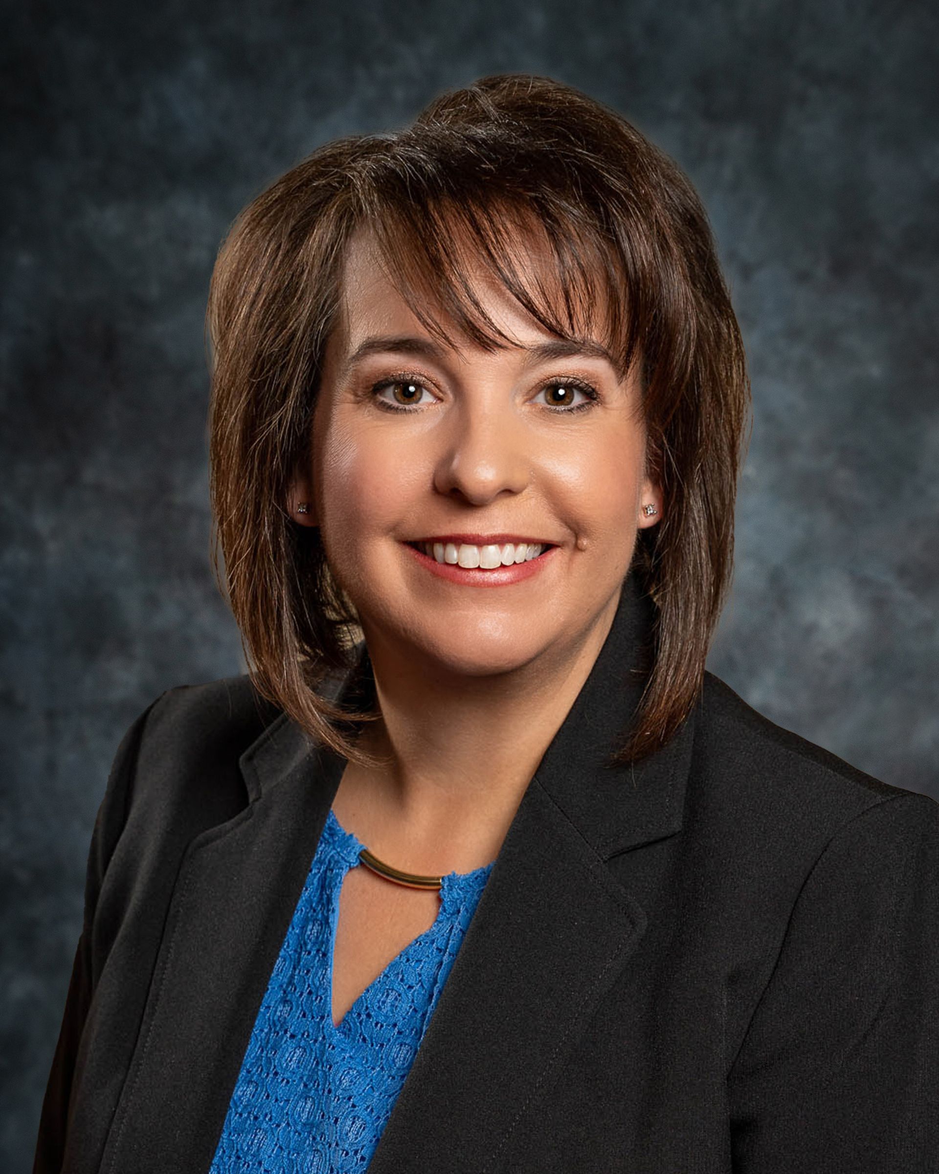 Woman with short brown hair smiles, wearing a blue blouse and black blazer.