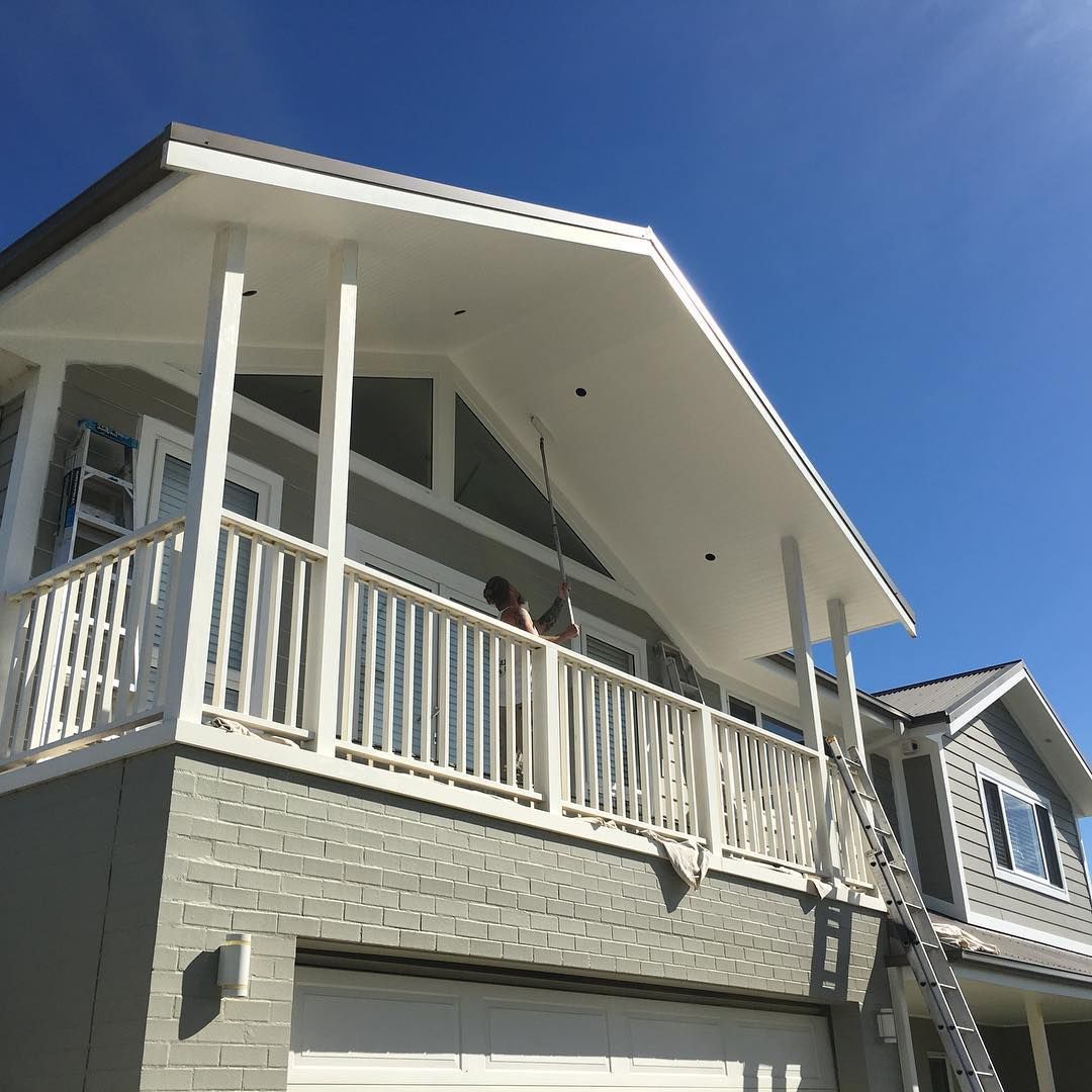 House with a white porch and railing, being painted gray against a clear blue sky.