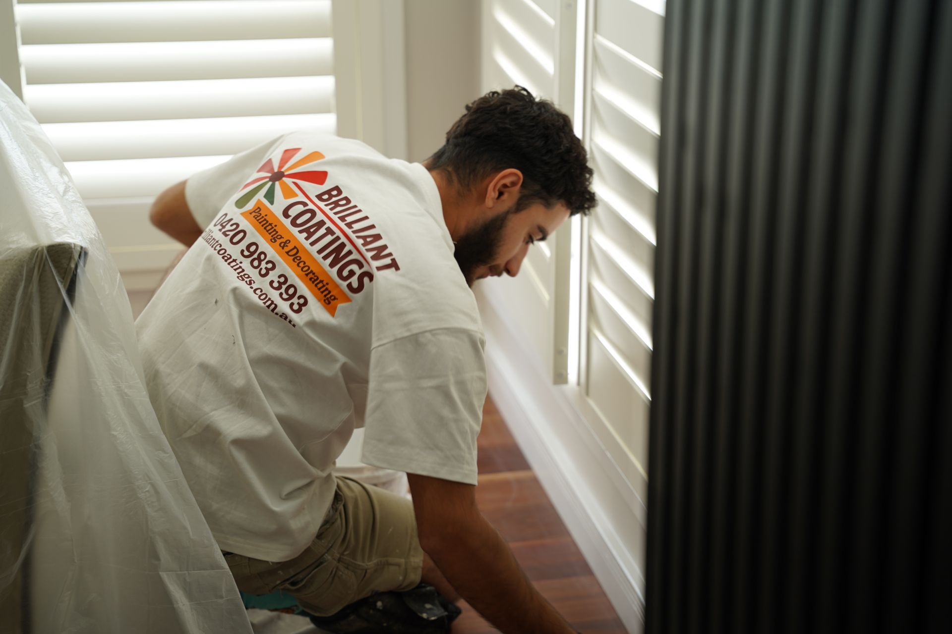 Man in a white shirt with logo painting trim near white shutters and a dark vertical panel.