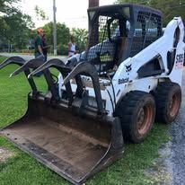 White skid-steer loader with a grappling attachment parked on grass near a person in the background