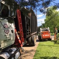Workers with a red ladder loading a truck and skid steer on a tree-lined driveway