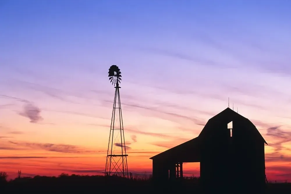 Silhouette of a barn and windmill at sunset under a pink-orange sky