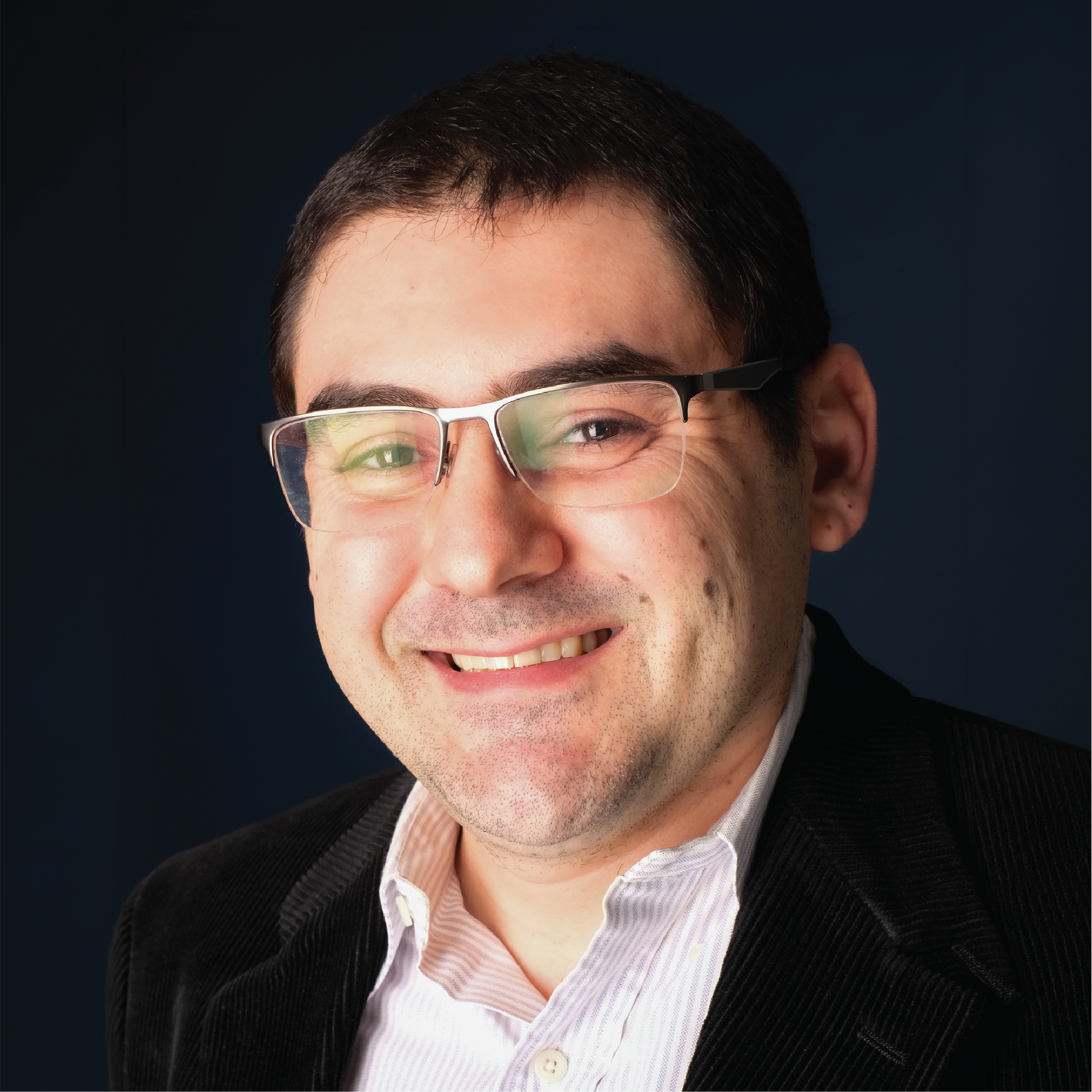 Headshot of a smiling man in glasses, white shirt, and striped tie against a blue studio backdrop