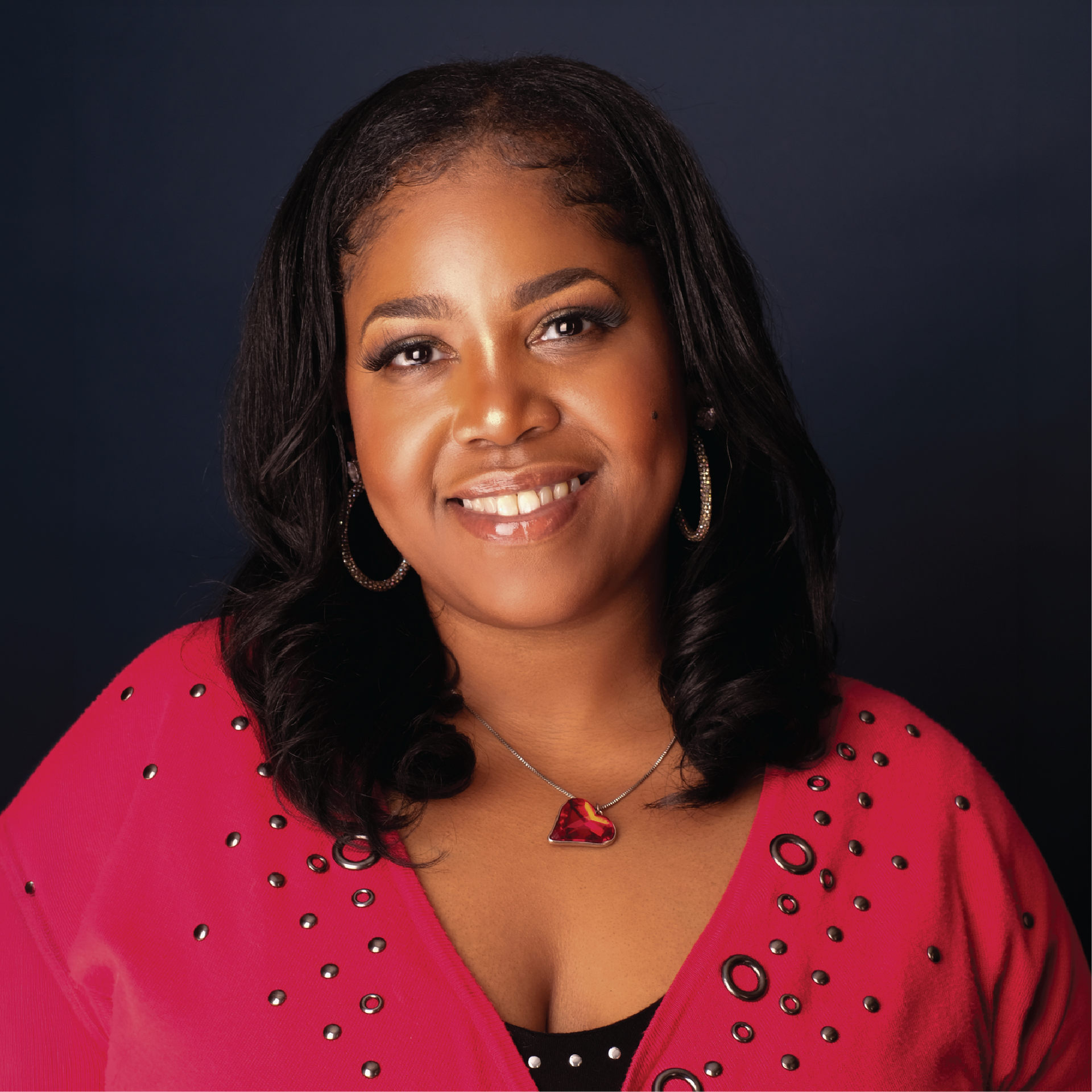 Smiling person in a pink top and red necklace against a dark studio background