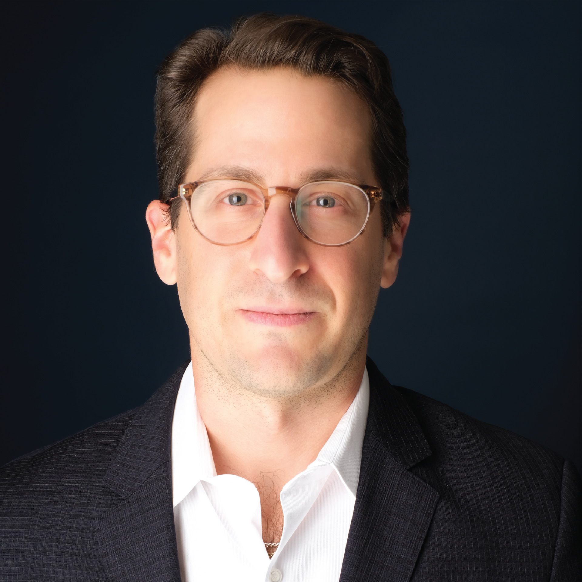 Studio portrait of a man in glasses, white shirt, and blue tie against a dark blue background