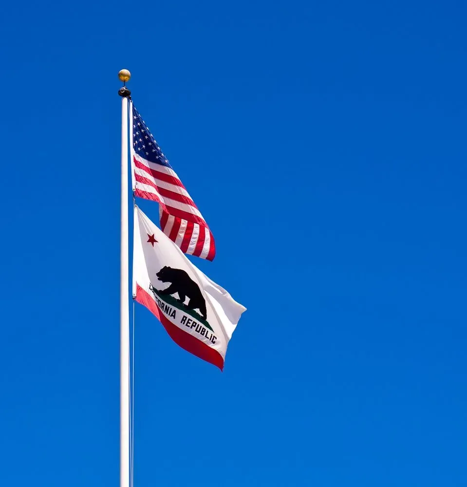 Two flags flying on a pole against a clear blue sky, including the U.S. and California flags