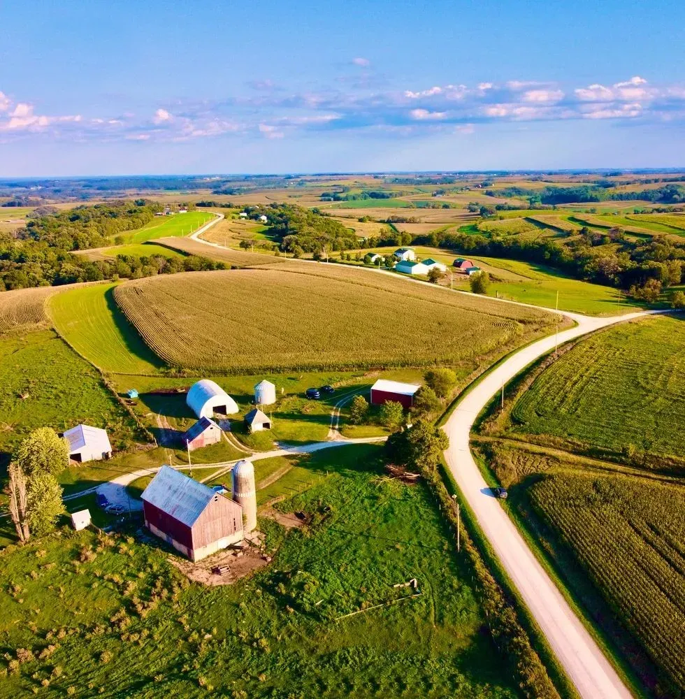 Aerial view of a rural farm with fields, barns, silos, and a winding road under a bright blue sky.