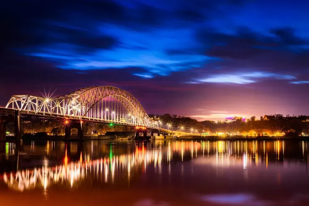 Illuminated bridge over a river at dusk, with colorful sky and shimmering reflections