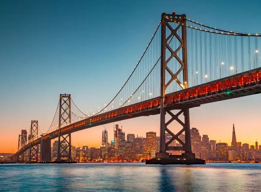 Suspension bridge at sunset over a city skyline and calm water, with warm lights glowing on the span