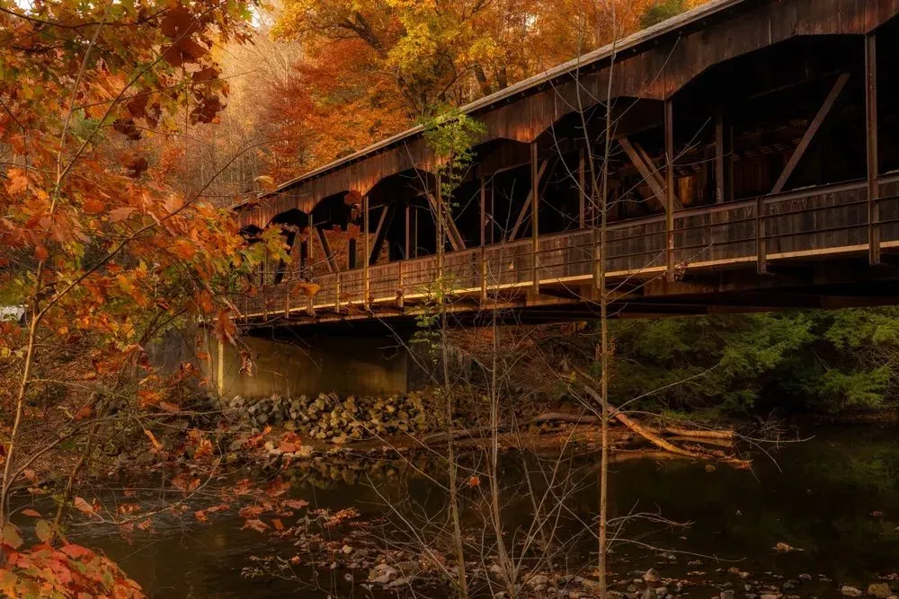 Covered wooden bridge over a calm river in autumn, with orange foliage reflecting in the water