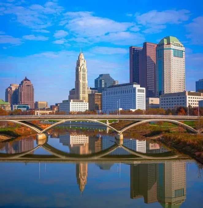City skyline with a bridge spanning a river, reflected in calm water under a blue sky.