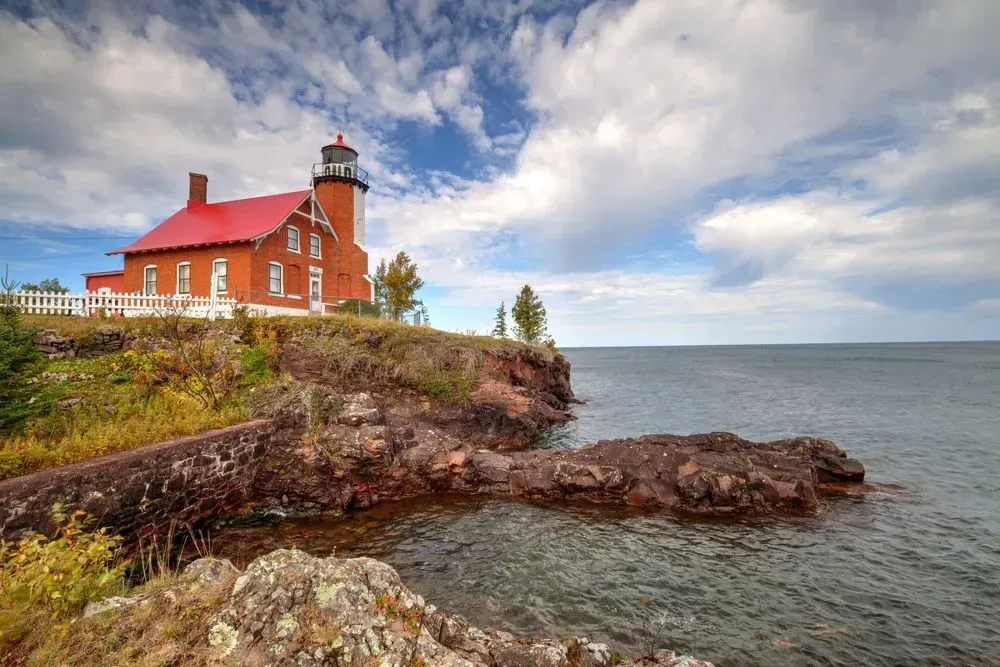Red-roofed lighthouse on a rocky cliff above a calm gray sea under a cloudy sky
